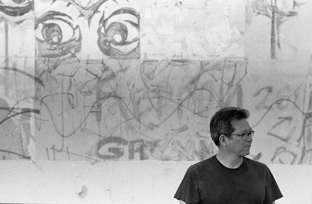 sesshu foster standing in a black t-shirt in front of a vandalized wall
